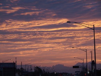 [The metal posts of the street lights are dark agains the purple-beige clouds in the sky. The sky is full of clouds, but at the horizon are dark purple shapes that might be mistaken for mountains but are actually thicker clouds at ground level.]